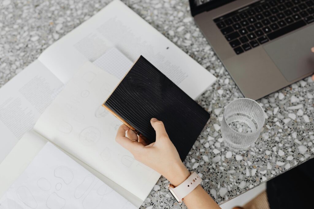 Overhead view of a hand holding a black notebook on a terrazzo table with a laptop and glass of water.